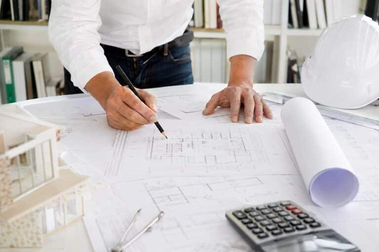 Maître d’œuvre étudiant des plans de construction sur une table avec casque de chantier, maquette architecturale et plans techniques dans un bureau lumineux